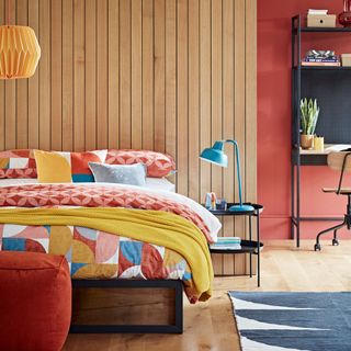 a bright bedroom with a red, blue, white and yellow geometrical pattern duvet cover on the bed, up against a wooden slat wall, with a red-orange wall next to it with a industrial style desk
