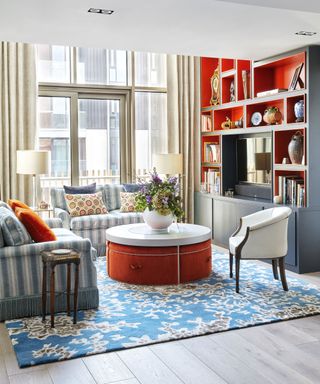 Living room with wooden flooring, blue and cream rug and sofas, dark wood and orange display shelves and french doors onto the balcony