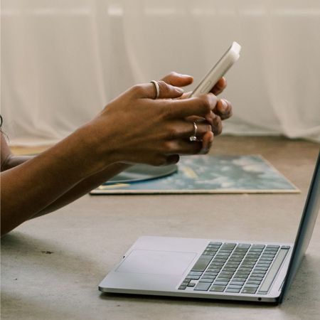 A woman scrolling on her phone, working on her laptop on the floor