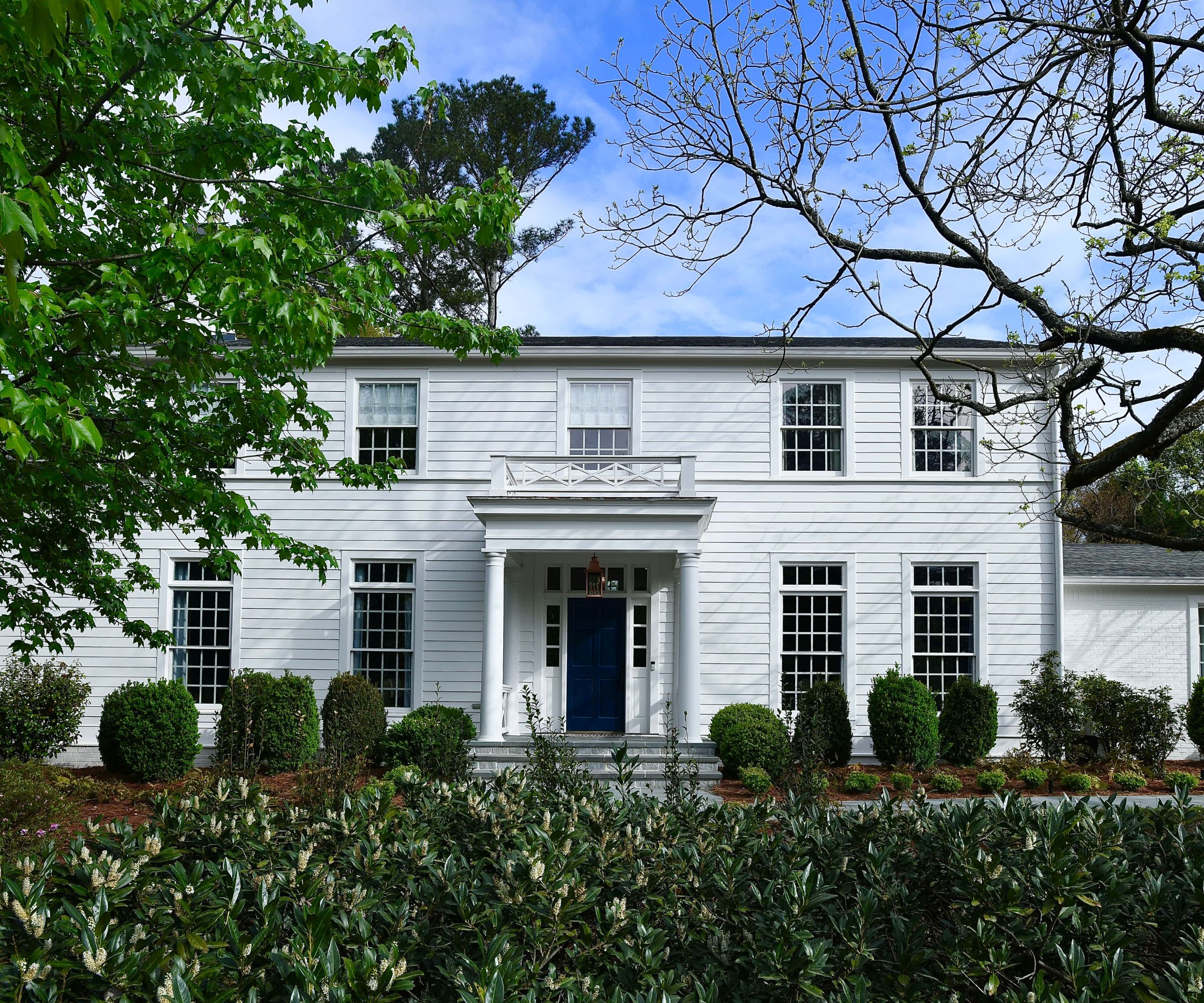 A large traditional home with a white exterior and Georgian-style windows. A dark blue front door with a porch with pillars and lots of greenery in the foreground.