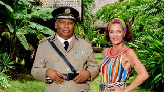 A posed shot of Commissioner Selwyn Patterson (Don Warrington) and Catherine Bordey (Élizabeth Bourgine) in front of some lush greenery. The Commissioner is in his work uniform, and Catherine is wearing a dress with bright multicoloured vertical stripes.