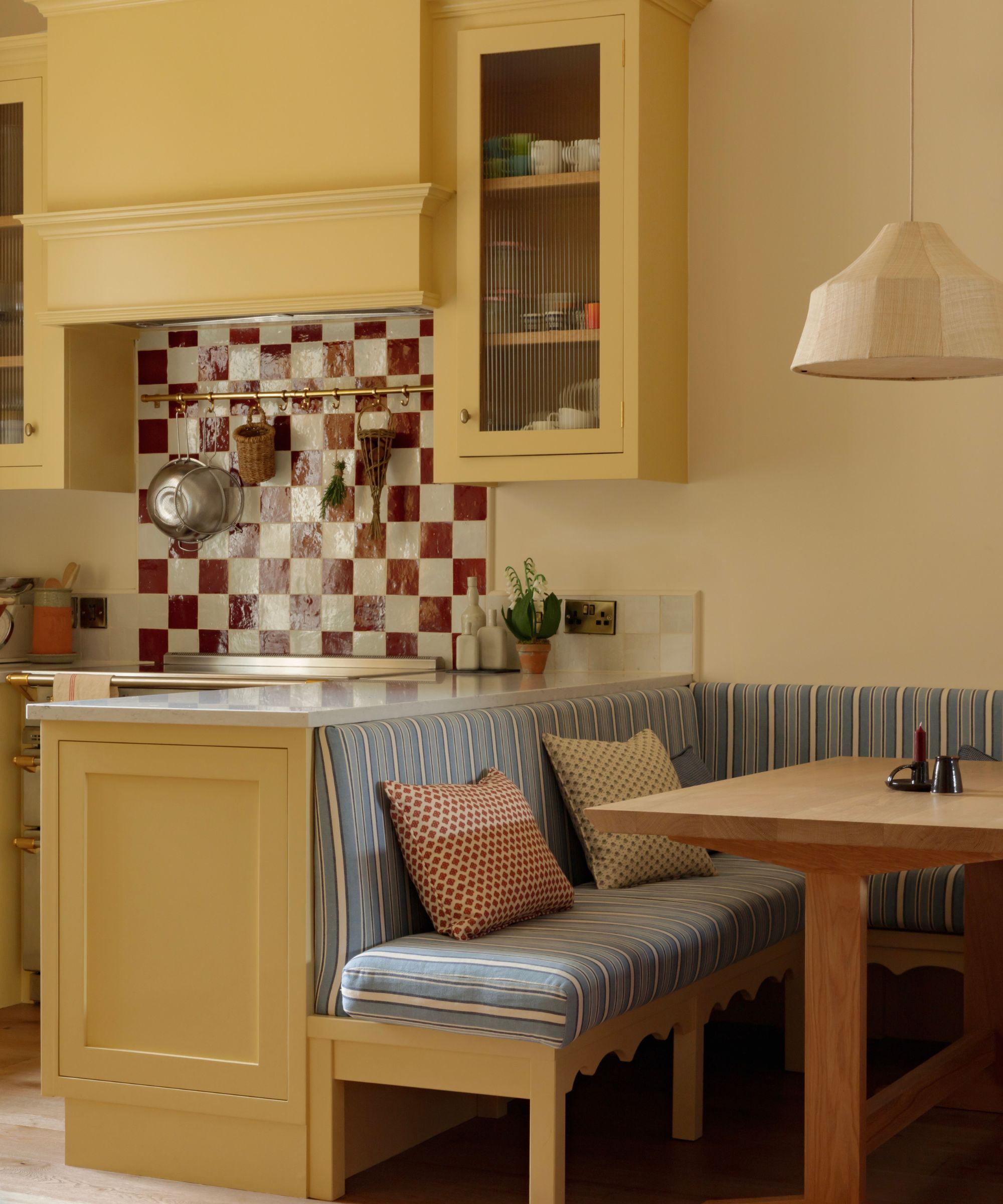 A cozy kitchen with butter yellow cabinets, red checkerboard backsplash tiles, striped blue banquette seating, and a wooden table.