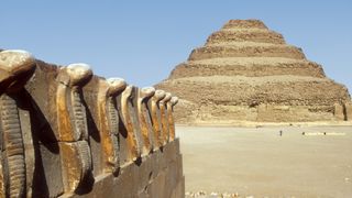 In the background we see the Step Pyramid in Saqqara, Egypt. It has 5 steps, each level higher getting shorter. In the foreground we see a row of stone cobra carvings on a wall.
