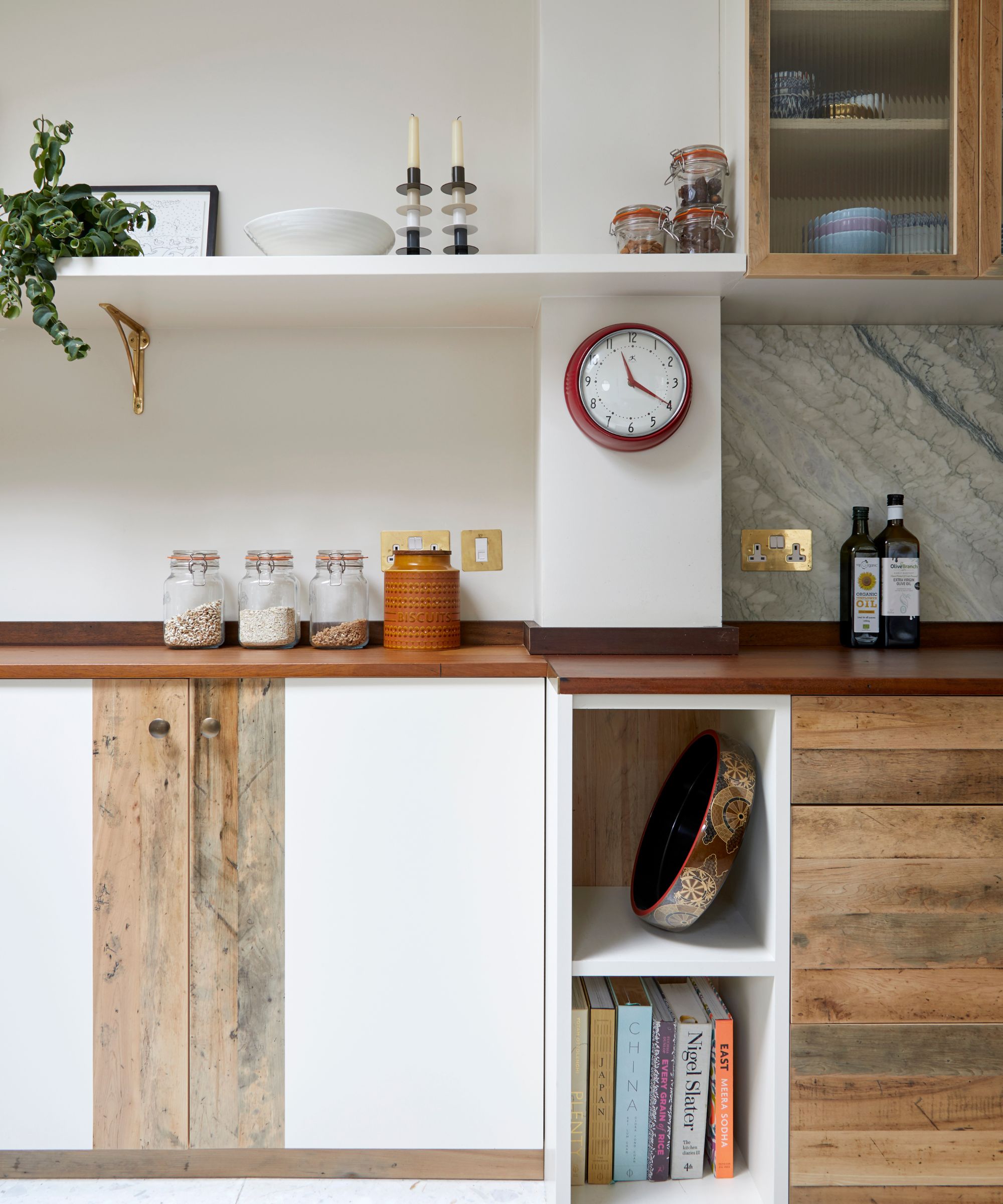 a kitchen with rustic wood countertops, wood cabinets, an open shelf with decor on it, a clock hanging on the wall, and grains in jars on the countertop