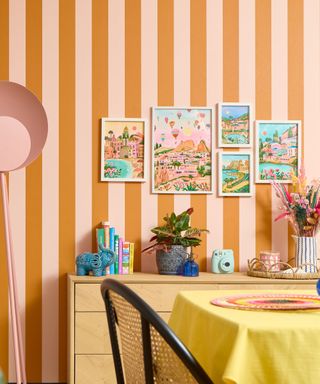 A dining area with peach and orange striped wallpaper with wall art prints on the walls, a wooden console table with plants on, and a yellow clothed dining table