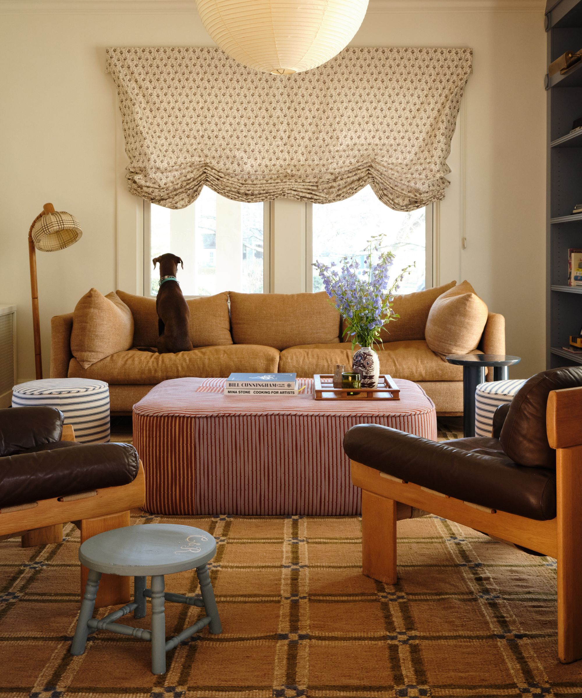 a soft living room with a large yellow check rug, striped ottoman, two leather and wooden armchairs, a dark yellow couch and paper lampshade