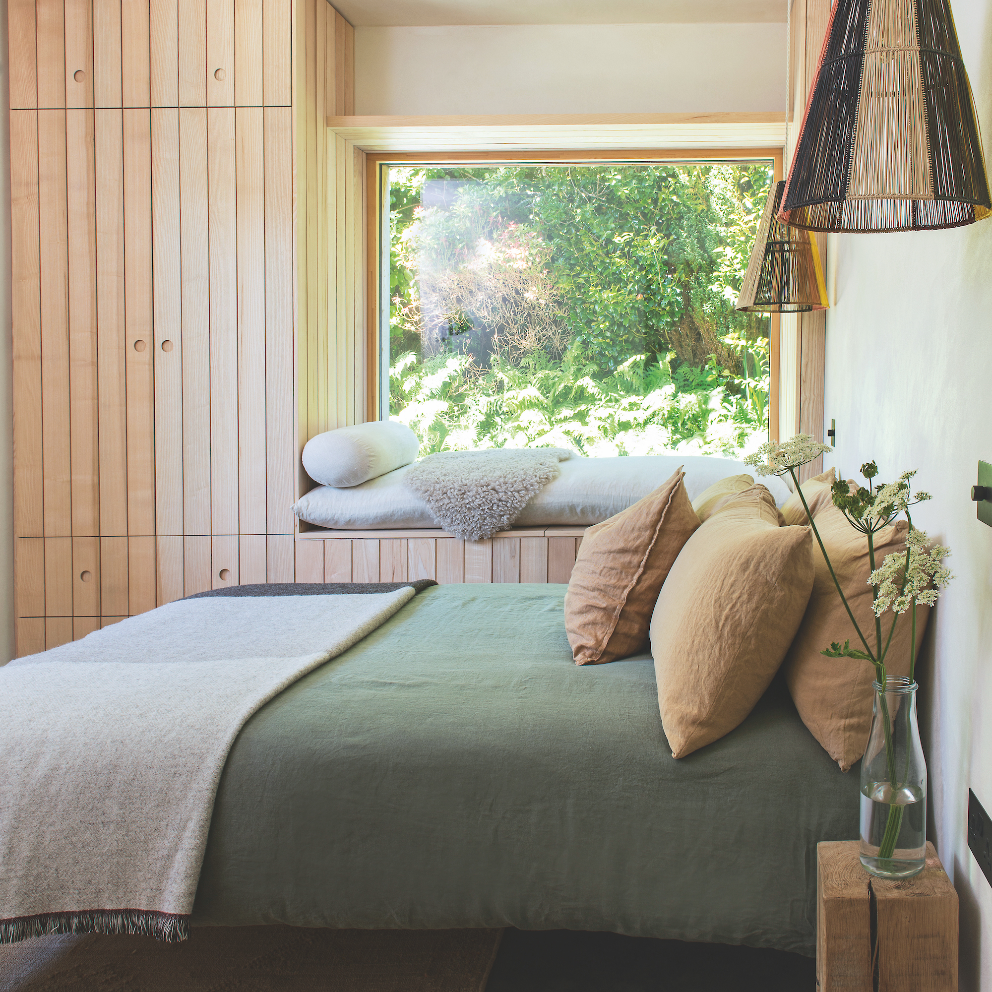 contemporary bedroom decorated in simple scandi style with built-in wooden wardrobes, large window overlooking garden with bench window seat and bed dressed with green linen