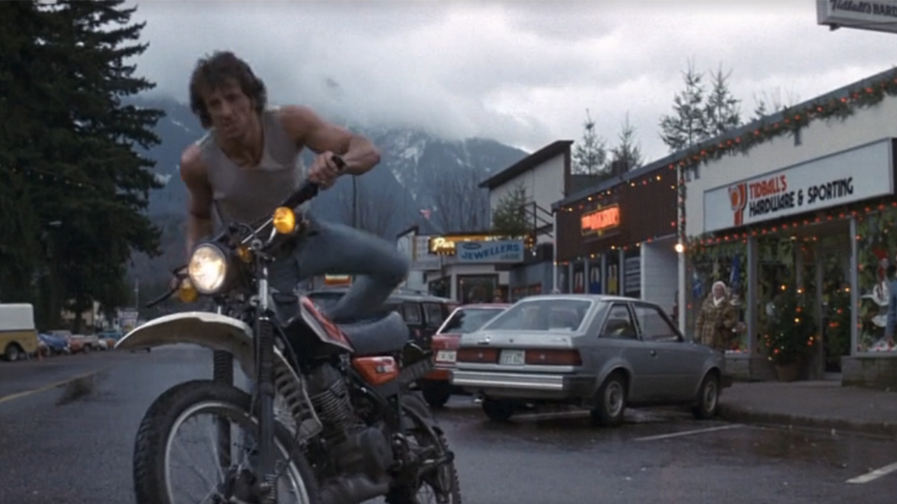 Sylvester Stallone getting on a motorcycle in front of stores with Christmas lights up in First Blood.