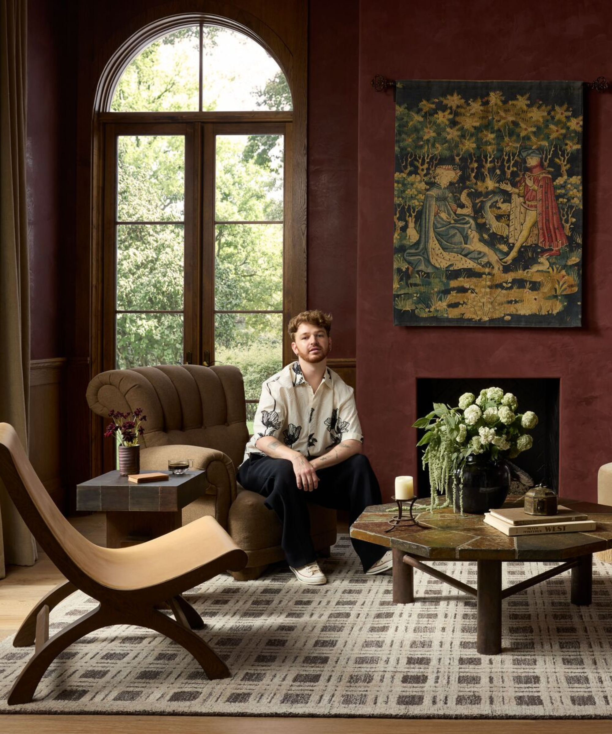 a living room with dark red paint, a medieval-inspired tapestry, a checker printed rug, two accent chairs, a geometric coffee table, and a man sitting on one of the chairs