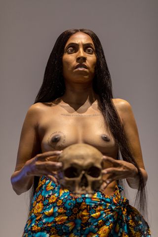 The naked torso and head of a dark-skinned female with her hands holding onto the head of a skull.