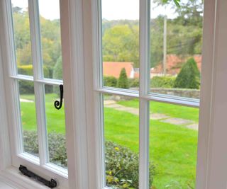 A close-up shot of a new traditional-style timber casement windows by The Sash Window Workshop. The frames and mullions are painted in white and one window features black, traditional-style window furniture. The windows provide a view of a larger, rural garden.