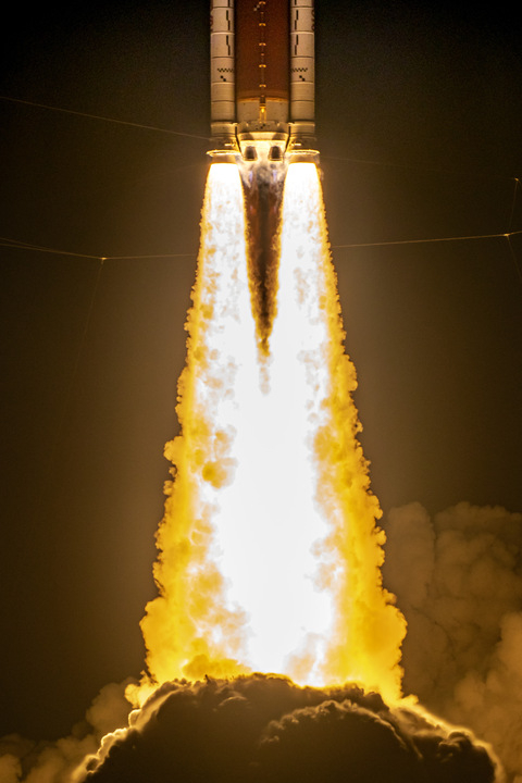 the bottom half of an orange rocket with two solid rocket boosters is seen with flames erupting from its engines. The image is filled with the fiery plume, based at the bottom in smoke.