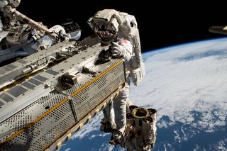 astronaut grasping on to a piece of space equipment while mounted to a robotic arm. the earth is behind