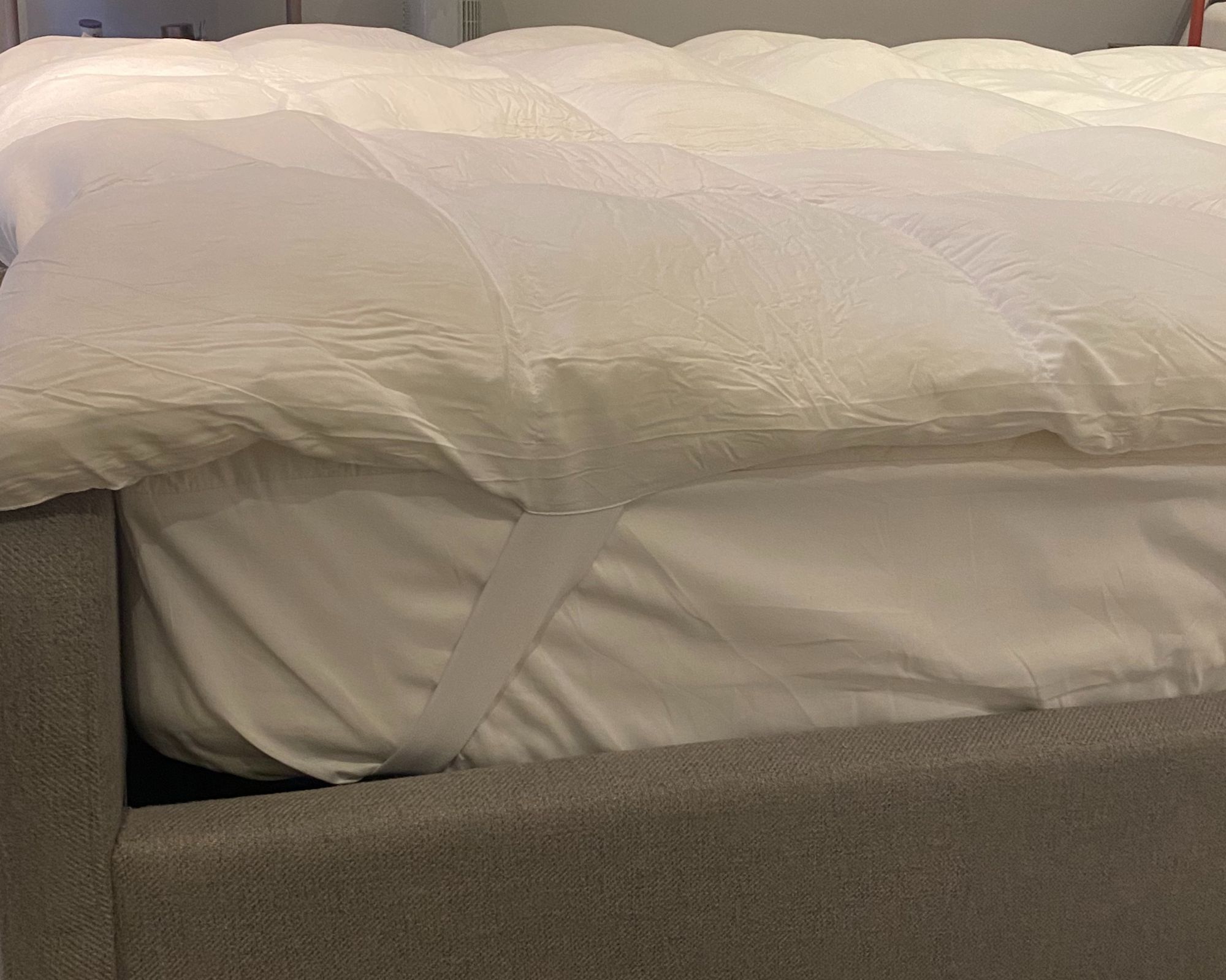 A white Coop Home Goods retreat mattress topper shown from the side on a light grey bed frame.