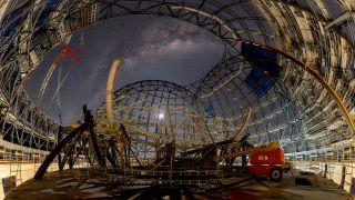 an extremely wide view staring up at the night sky as the milky way looms overhead. scaffolding and the bones of a large domed structure surround, but for a hole in the ceiling for the stars.