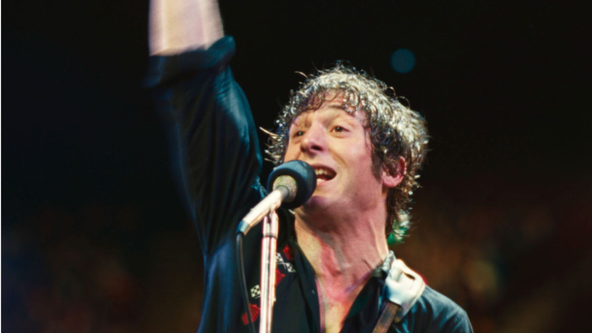 an image of jeremy allen white playing bruce springsteen in the movie Deliver me from Nowhere. He is performing on stage during a hot evening in a black shirt and playing guitar &amp; singing