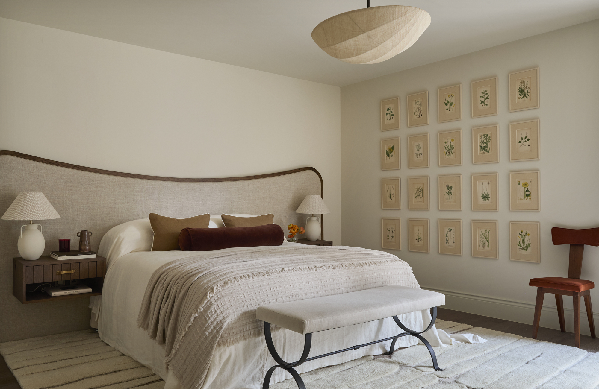 A bedroom with taupe walls, a large headboard with brown trim, and a gallery wall of botanical paintings