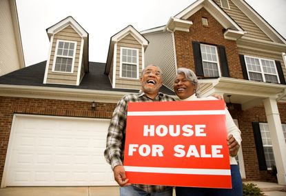 A couple stands outside of their house with a for sale sign
