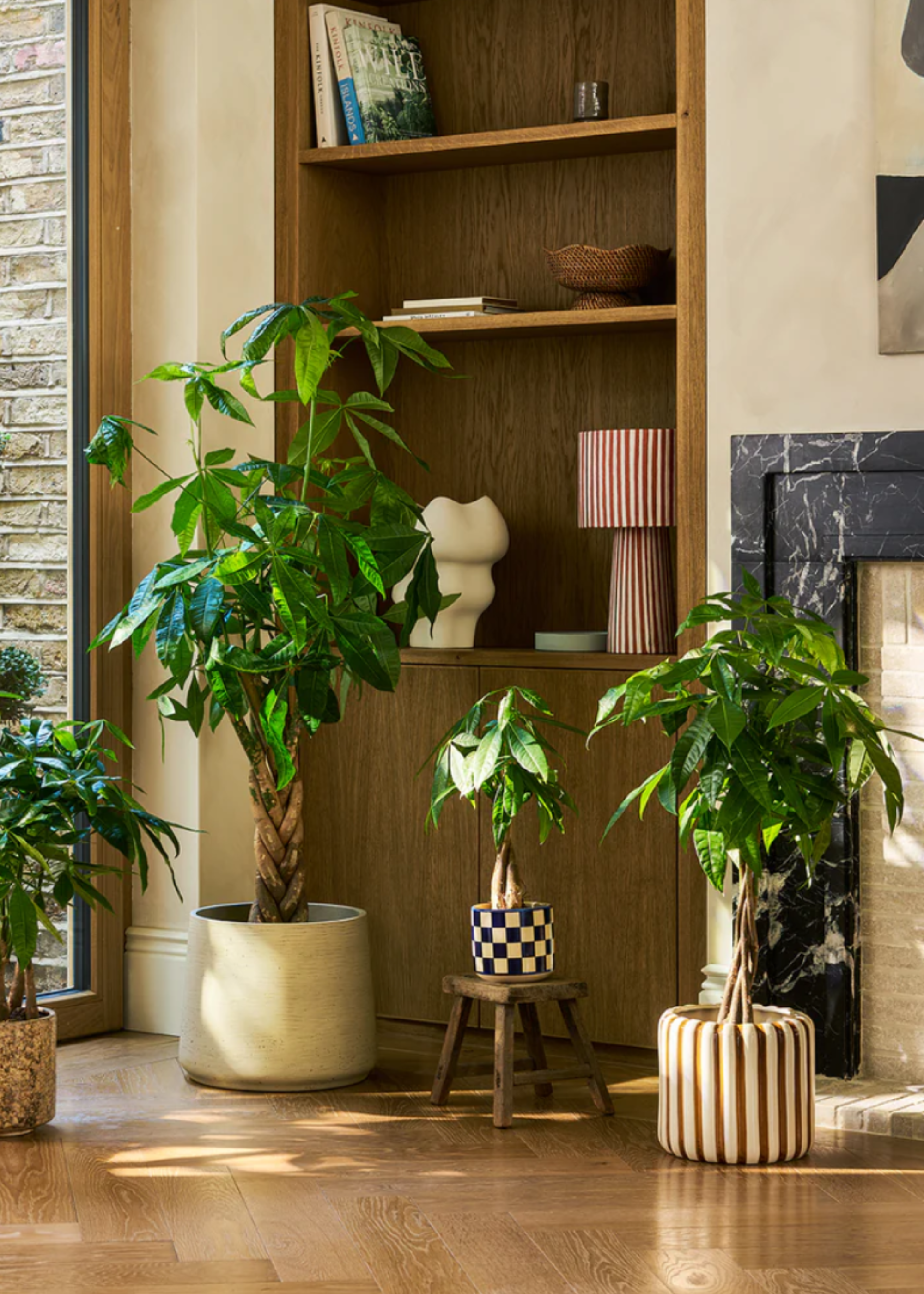 A minimalist living room corner with potted paper plants in striped and checkerboard planters by a stone fireplace and a wooden shelving unit with a striped lamp, a woven bowl, a ceramic vase and a couple of books