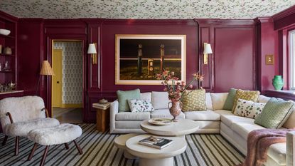 A living room with burgundy paneled walls in a gloss finish and a floral wallpapered ceiling. A striped carpet, a large cream sectional with green, yellow, and floral cushions, a modern three-piece coffee table, artwork on the wall with two sconces, and a shearling accent chair.