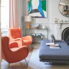 Cat sitting on large upholstered footstool in living room with orange accent chairs and colourful artwork