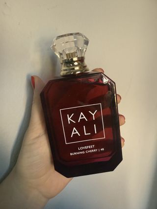 Brooke knappenberger hand with red nail polish holding up Kayali Lovefest Burning Cherry perfume bottle against grey wall