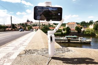 The Hohem iSteady v3 gimbal in white sits on a ledge. It is being used as a tripod to record video footage via a mounted iPhone.