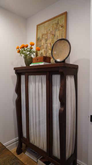 Image of the corner of a room. There is a vintage, dark wooden cupboard with glass-panelled doors. There are white sash curtains installed, and a yellow artwork and orange flowers on top of the cupboard.