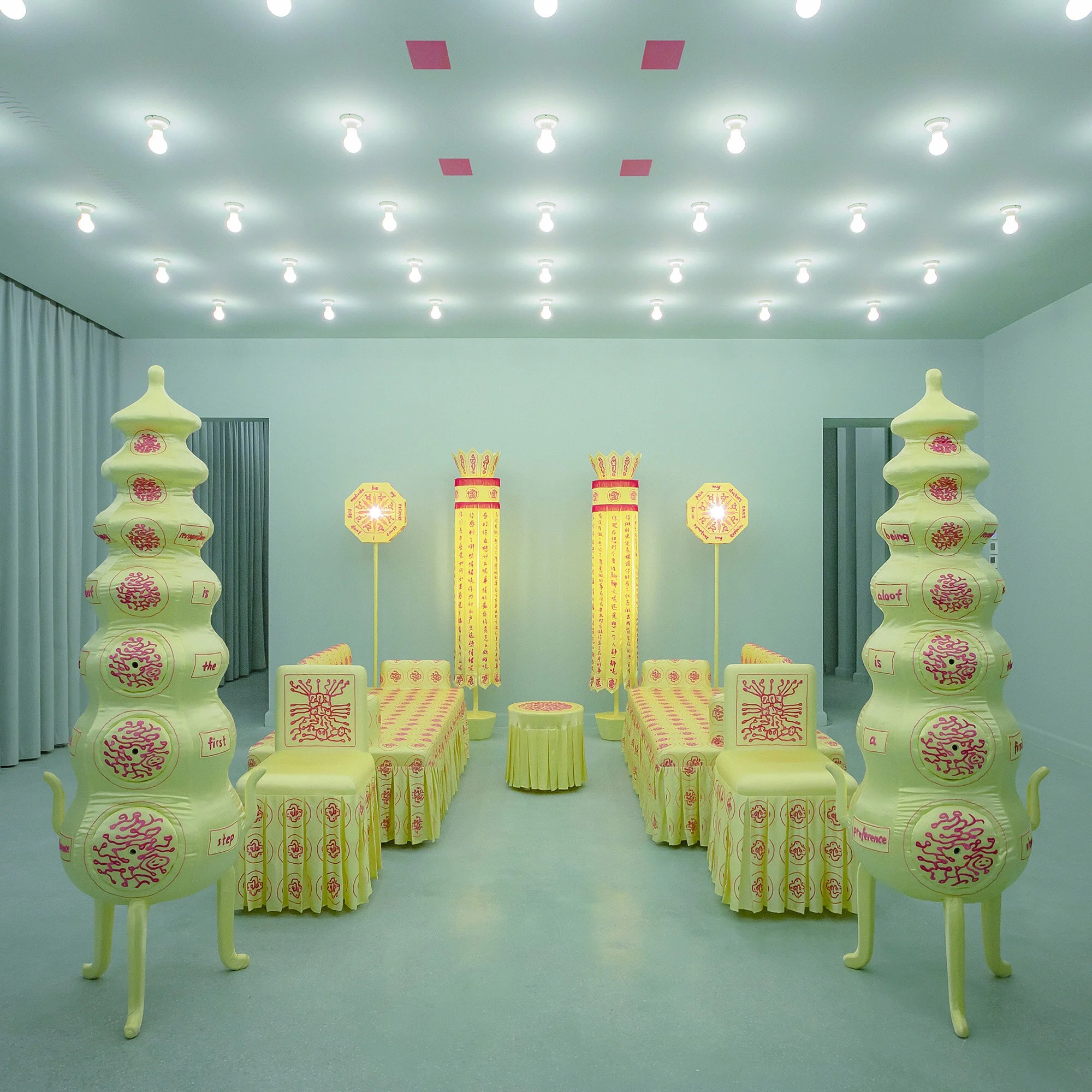 A mint green-shaded room brought to life by old-style light bulbs used as sky light houses a selection of Chinese heritage-inspired furniture with a whimsical shape, including chairs, floor lamps, and more, in a muted neon yellow color, with hand-drawn inscriptions as well as drawing on them.