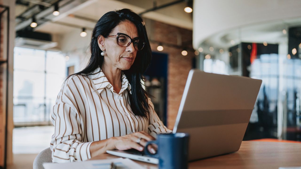 A woman with long brown hair, wearing thick-rimmed glasses and a button-up blouse, works on a laptop at a wooden table with a mug. 