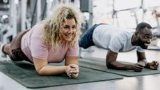 A woman and man perform the plank isometric exercise in a gym on mats. They are low to the ground, holding themselves up on their forearms and toes, their bodies elevated above the floor. Behind them we see the blurred outline of gym machines. 