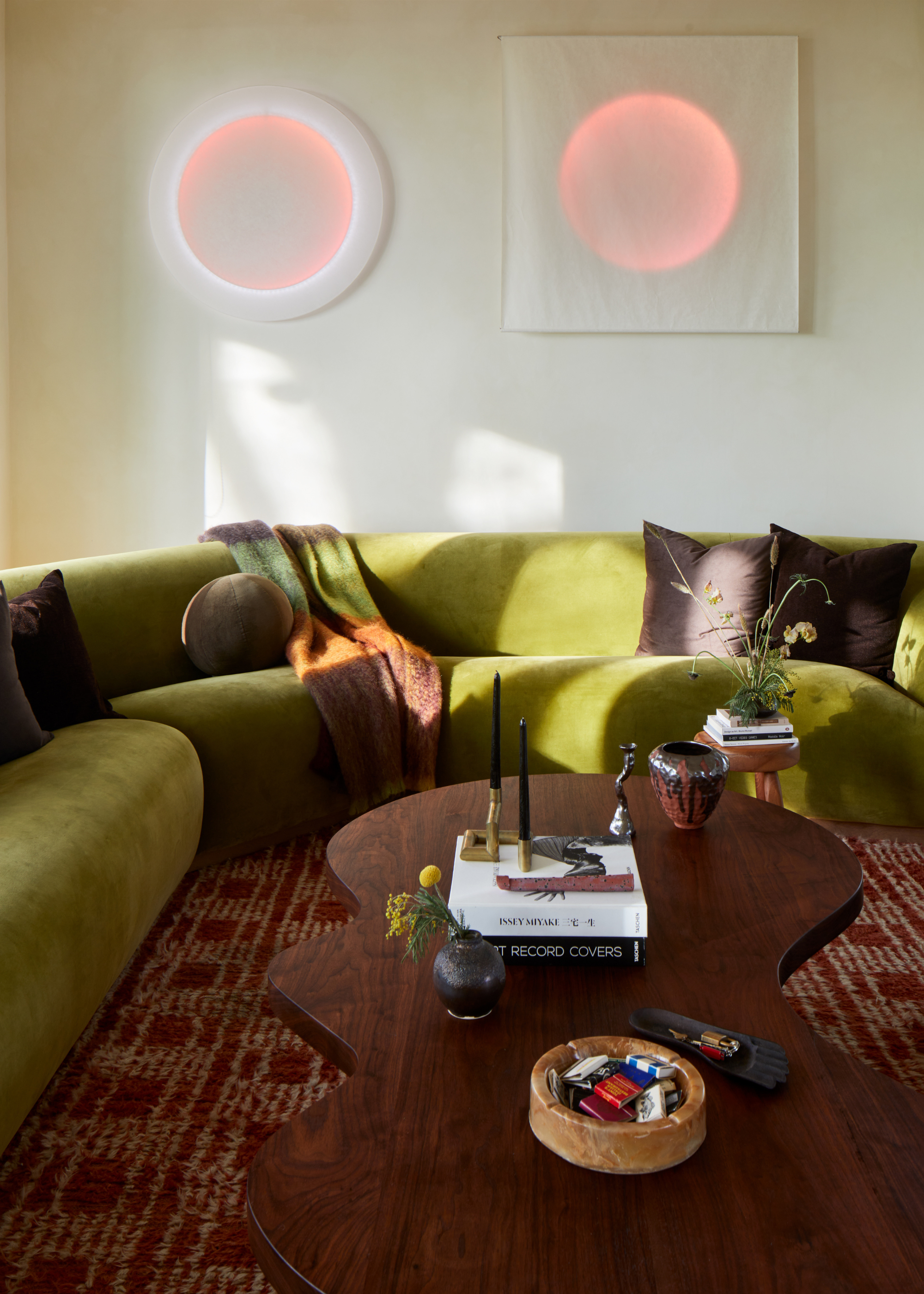 A white living room with white and neon wall features, a curved olive green couch with brown pillows, a wavy wooden coffee table, and a patterned rug