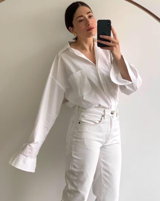 British fashion influencer Lucy Alston poses for a mirror selfie wearing a white button-down shirt, yellow-gold earrings, and white jeans.