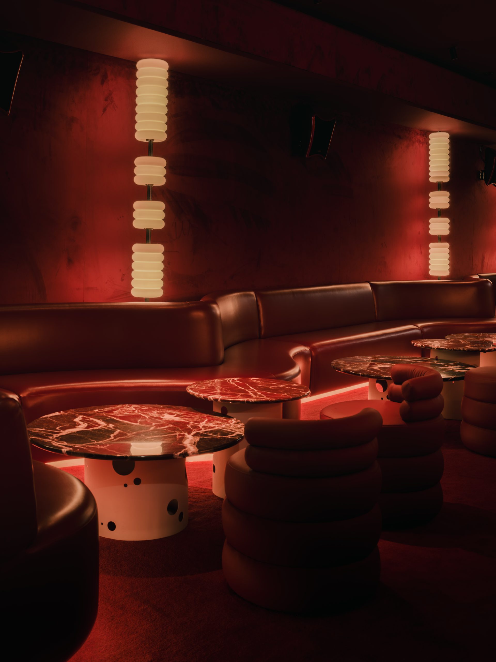 A dimly lit bar with red and orange-tinted interiors sculpted in wood, chrome, lacquer, velvet, and marble features sinuous banquettes, atmospheric sconces, geometrical skylights, and refined details.