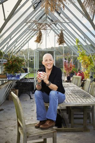 Sarah Beeny sits on the table in her potting shed, with her feet up on a chair, clutching a mug of tea. She is bald in this picture.