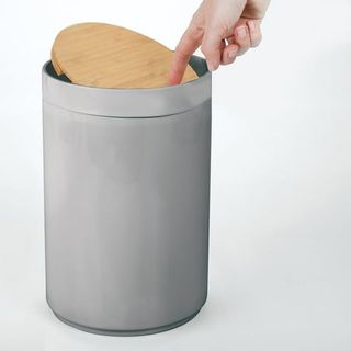 A grey plastic cylindrical bin with a bamboo swing lid. Someone's finger holding it open