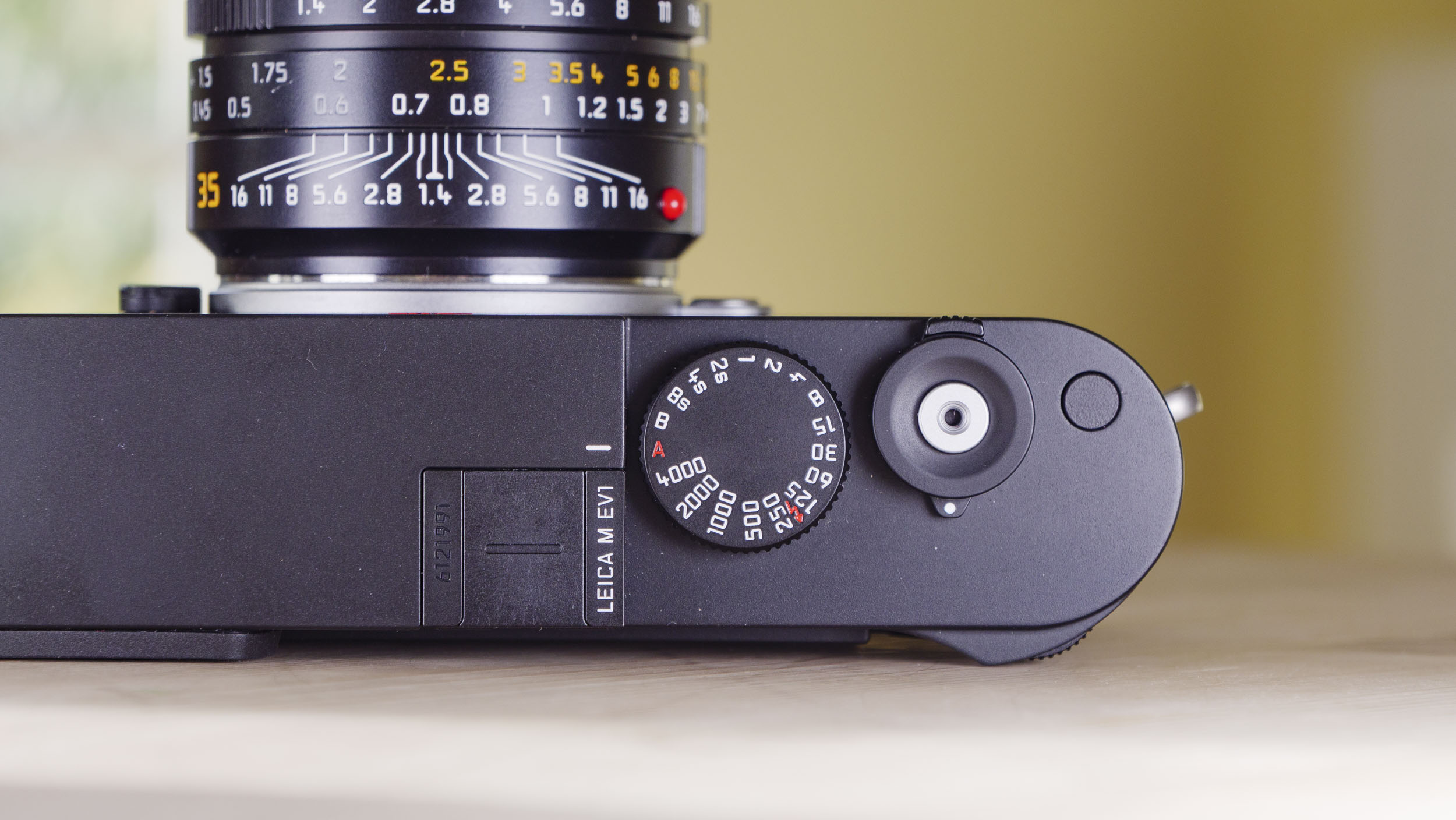 Top plate of the Leica M EV1 camera, which is resting on a wooden table with glass-panelled door backdrop