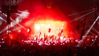 A night photograph of Glastonbury's Pyramid Stage lit up in red, with the crowd waving flags.