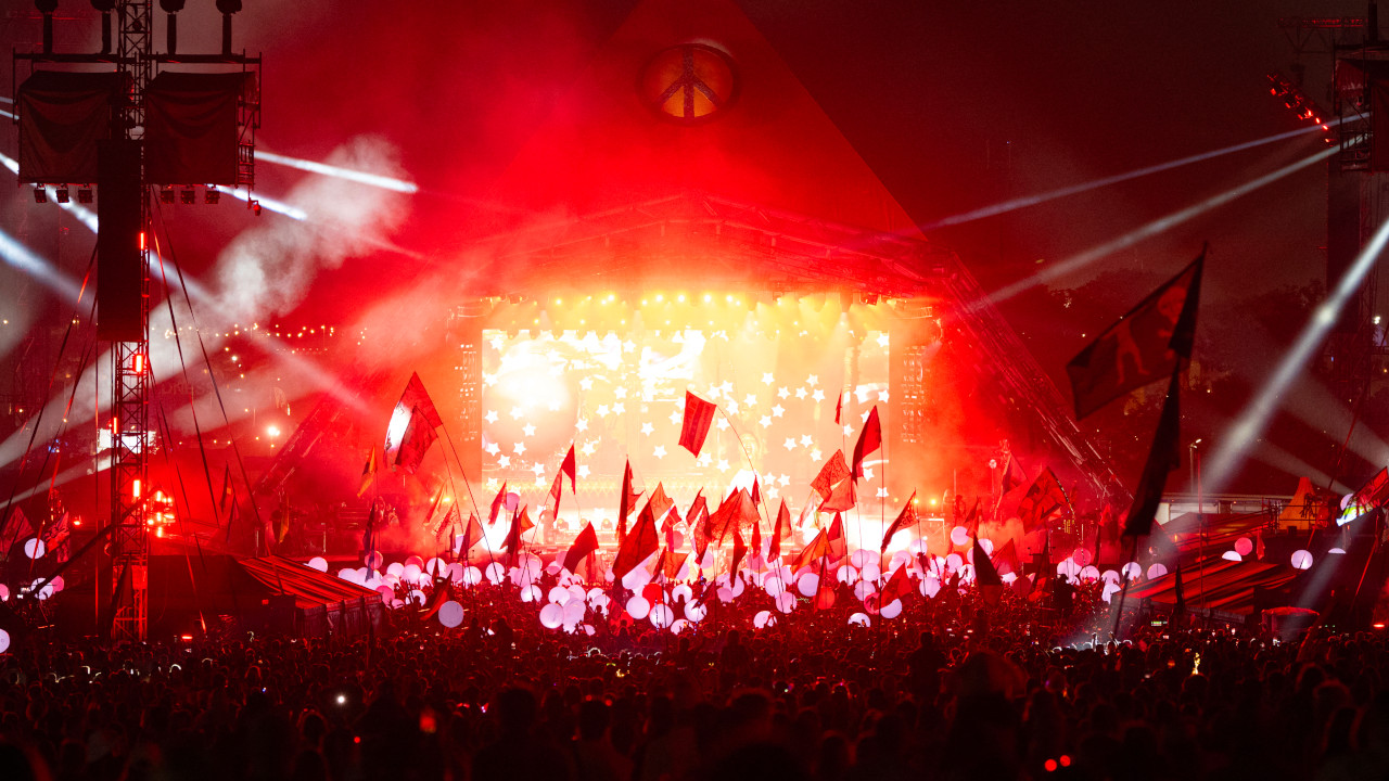 A night photograph of Glastonbury&#039;s Pyramid Stage lit up in red, with the crowd waving flags.