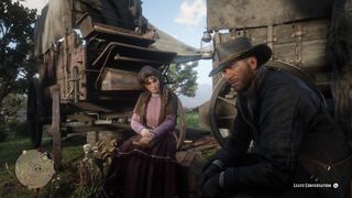 Red Dead Redemption 2 - Two people sit at a campsite in front of wagons