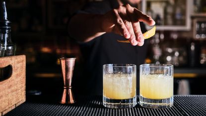 moody side shot of a bartender expressing citrus peel over two cocktails in highball glasses