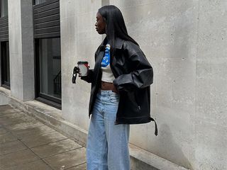 Woman wears crop top, baggy light-wash jeans, and sneakers with an oversize leather jacket while holding a disposable coffee cup.