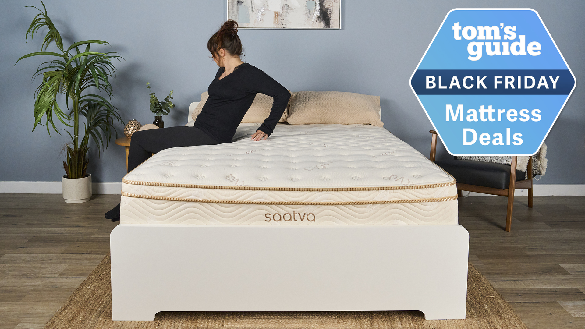 This image shows a member of our sleep team, Lauren Jeffries, sitting on the side of the Saatva Classic mattress on a bed frame in our Sleep Lab. In the top right corner is a Tom&#039;s Guide Black Friday mattress deals graphic
