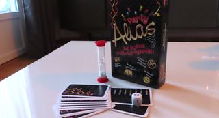 Ett party alias står uppställt på ett vitt bord med tillhörande spelkort utspridda framför tillsammans med en tärning och ett timglas.