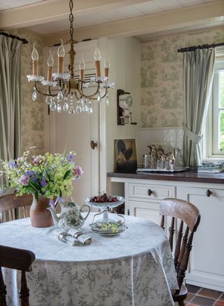round kitchen table with patterned cloth and green toile de Jouy wallpaper red tiles and chandelier