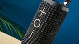 a black oblong-shaped bluetooth speaker with mesh covering and a carry strap photographed on a beige table in front of a navy wall