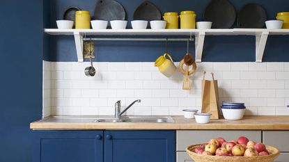 Blue painted kitchen with white tiled splashback and a white shelf above with a mix of yellow and white ceramics