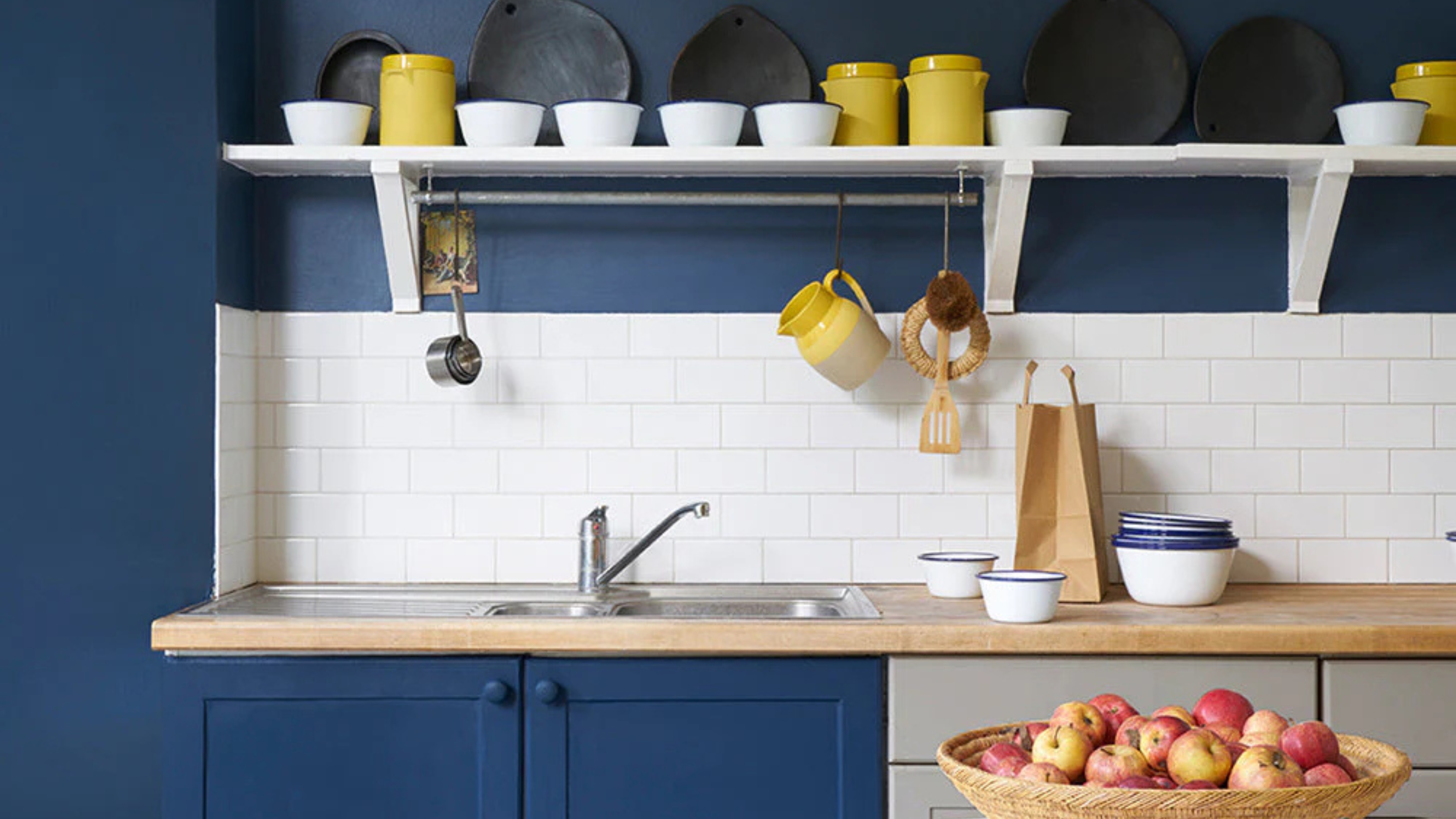Blue painted kitchen with white tiled splashback and a white shelf above with a mix of yellow and white ceramics