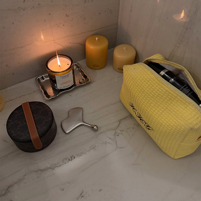 A yellow toiletry bag, stainless steel gua sha, and a few candles are pictured don top of a table or counter. 