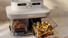 An air fryer with two glass baskets and a grey head unit on a marble kitchen counter. One basket contains meat, the other roasted vegetables